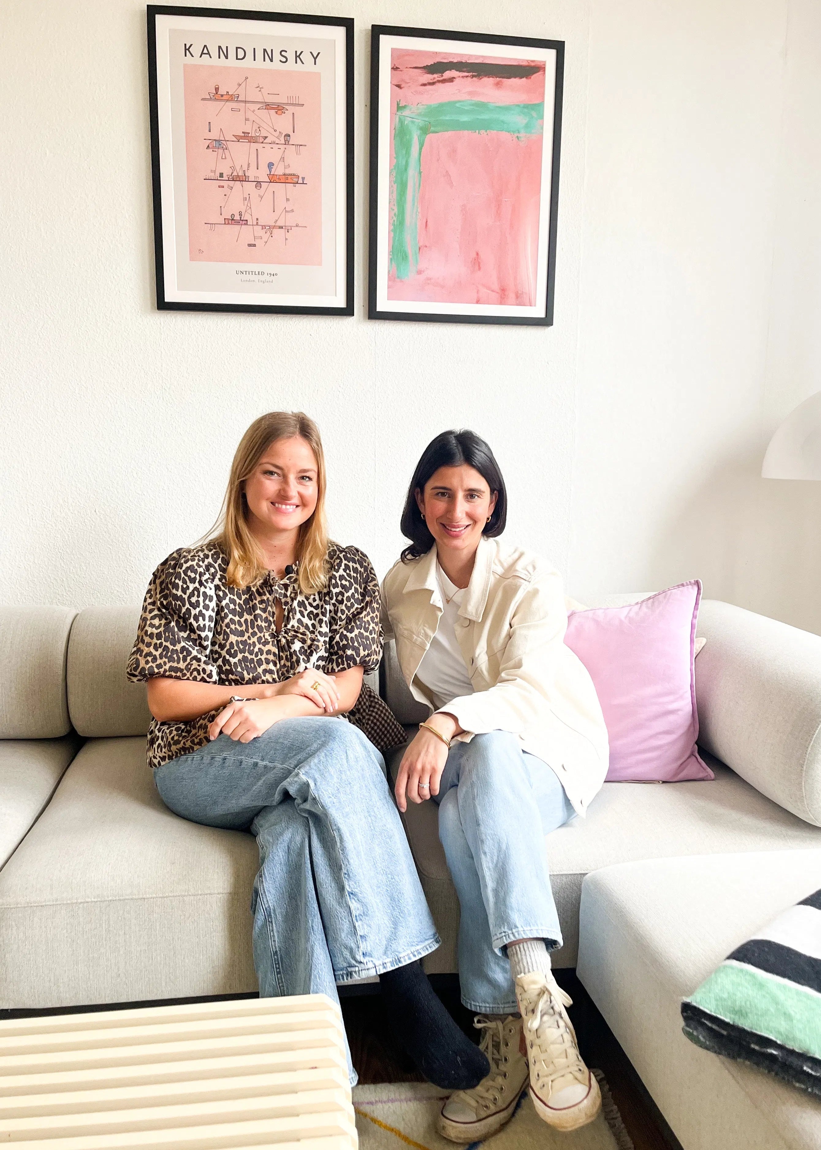 Two women sitting on a couch in a room with framed artwork on the wall.